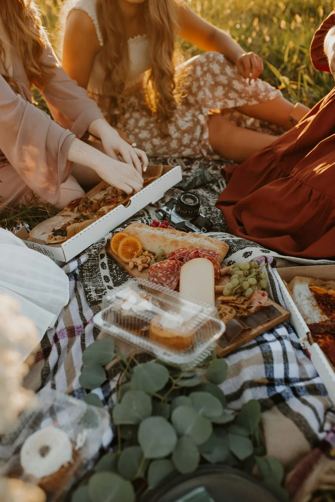 A golden-hour picnic spread with cheese, bread, grapes, and pizza on a blanket
